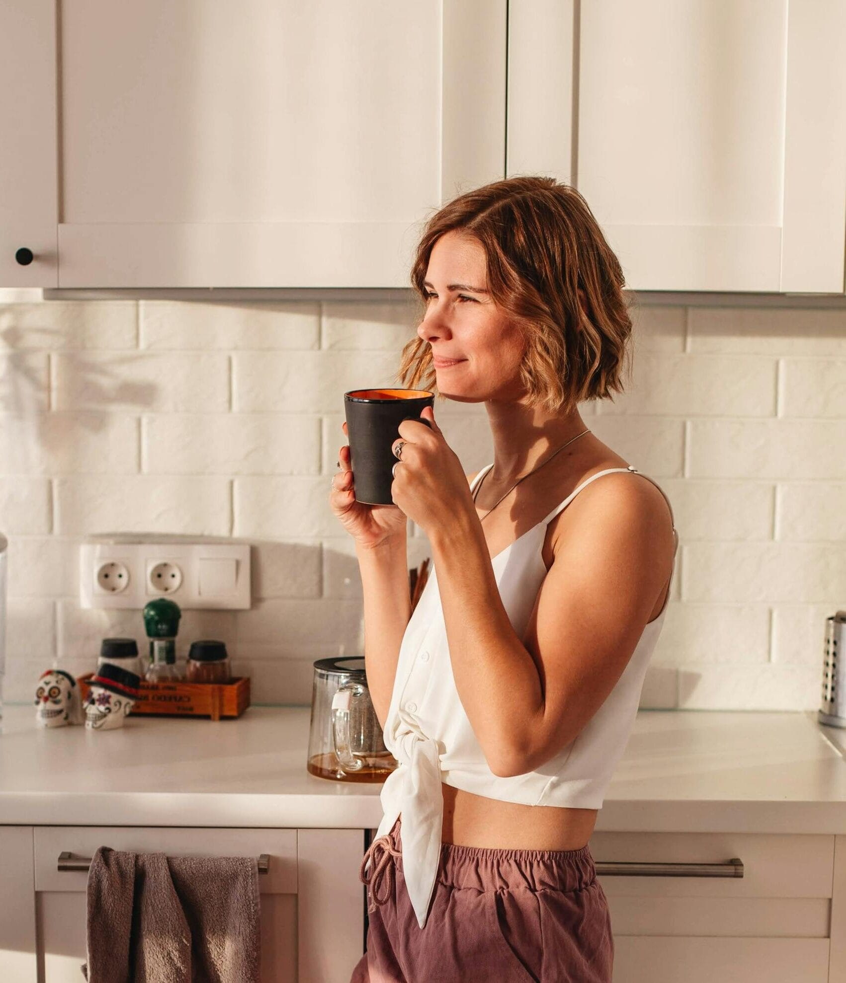 Start 3 Widok z boku uśmiechniętej kobiety w piżamie stojącej w jasnej czystej kuchni podczas porannego picia filiżanki świeżego napoju przy blacie i szafkach Photo by Anna Alexes